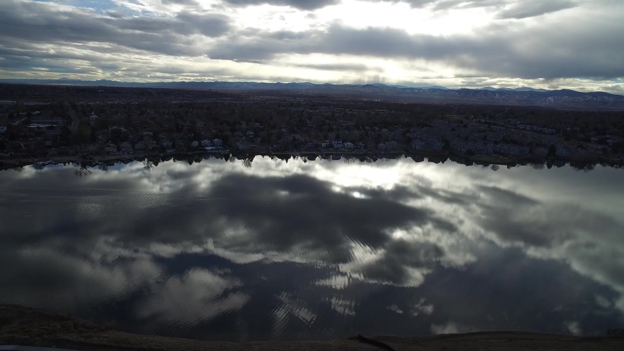 At 0:00, an aerial view in Westminster, Colorado looks across dark rippled water, bright cloud reflections, a low shoreline, distant homes, hills, and no visible people.