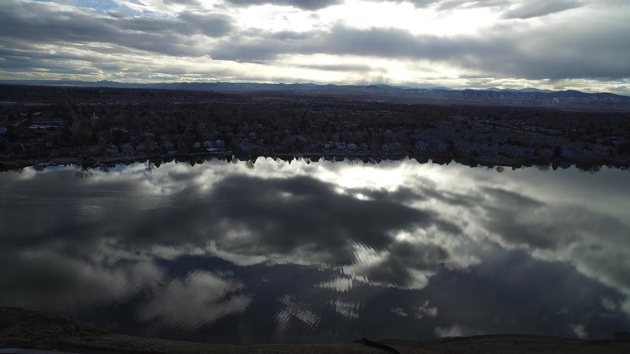 At 0:01, the Westminster, Colorado water view shows silver cloud reflections spread across the rippled surface, with a dark shoreline band and no visible people.