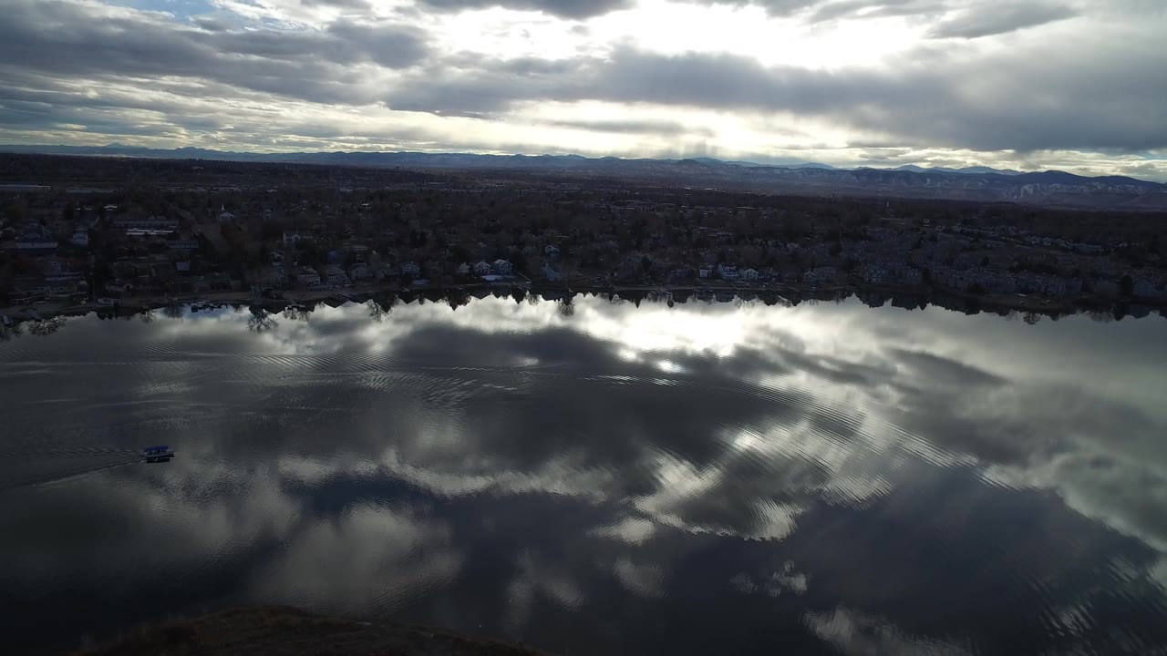 At 0:01, the small boat sits lower in the reflective water while the Westminster, Colorado shoreline and distant mountain ridge remain under layered clouds, with no visible people.