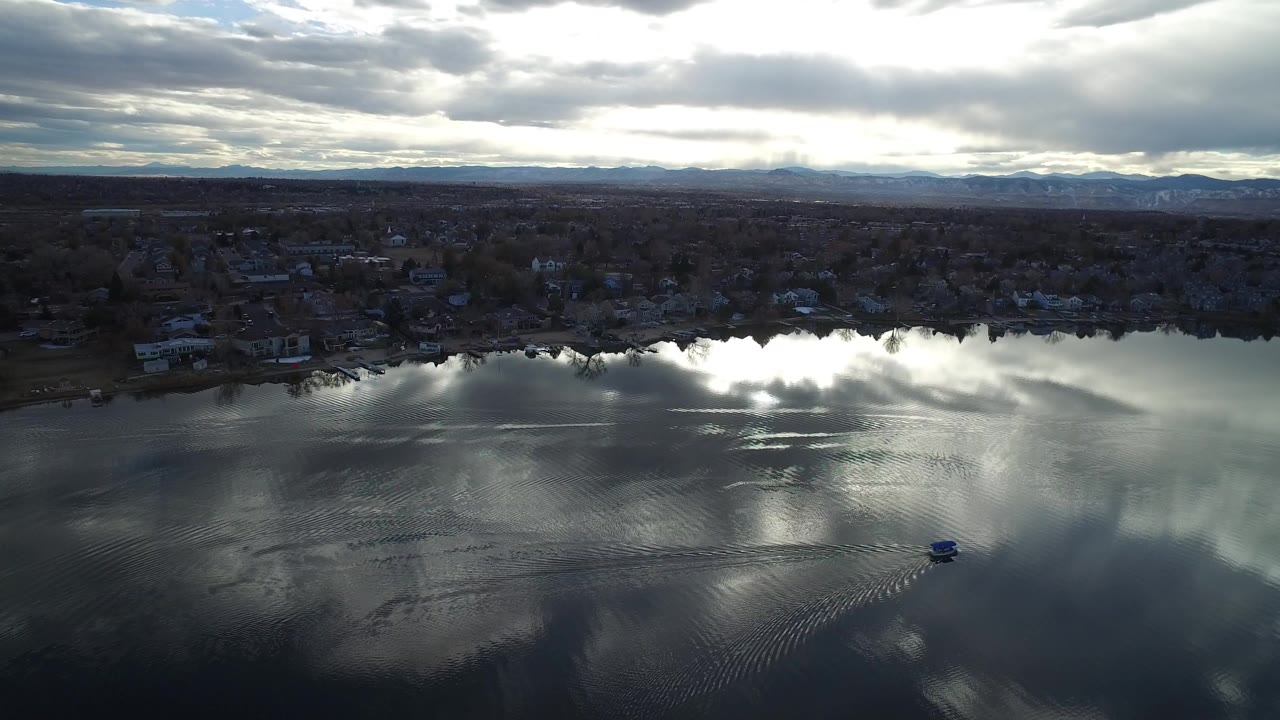 Aerial view of a lake, shoreline homes, cloud reflections, and distant mountains.