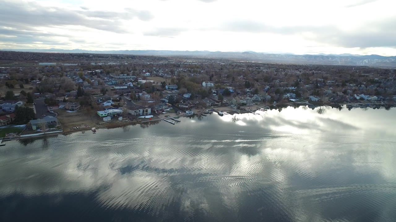 Westminster, Colorado aerial frame with bright cloud reflections on rippled water, lakeside houses and docks along the shore, distant suburbs, and no visible people.