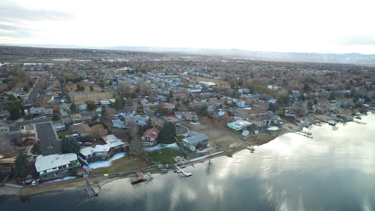 High aerial view over Westminster, Colorado homes beside reflective water, with docks, streets, leafless trees, distant hills, and no visible people.