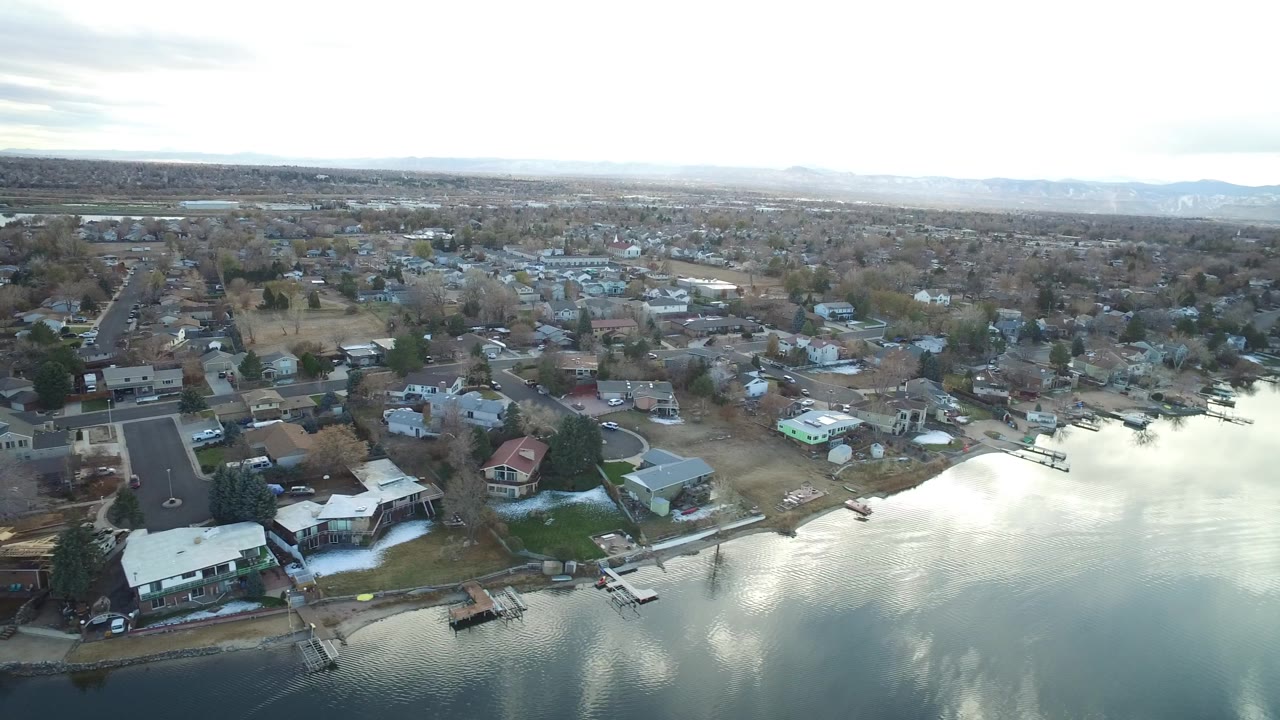 Aerial frame in Westminster, Colorado looking across calm water toward homes, small docks, neighborhood roads, bare trees, and no visible people.