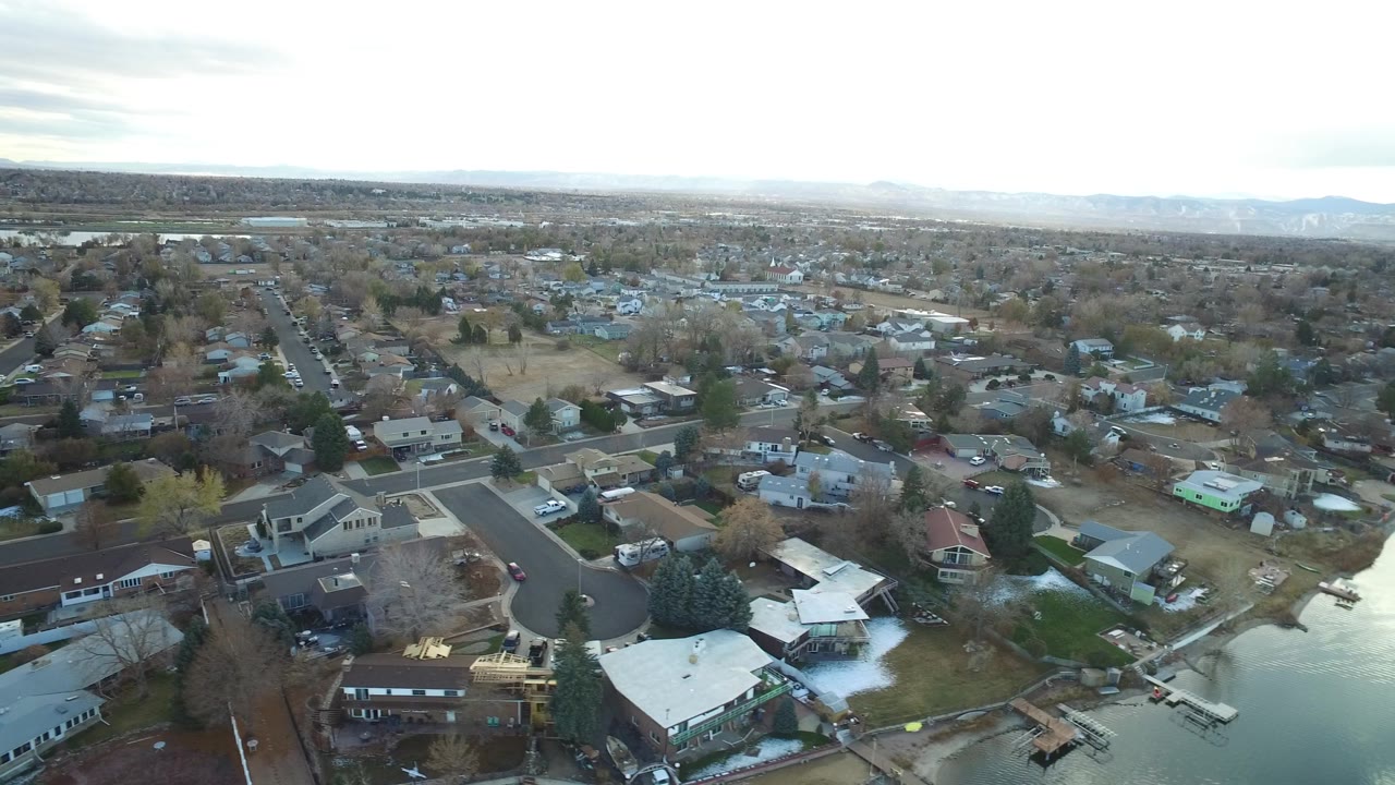 Aerial frame looking across Westminster, Colorado rooftops, fenced yards, a curved road, parked vehicles, and a bright clouded horizon, with no visible people.