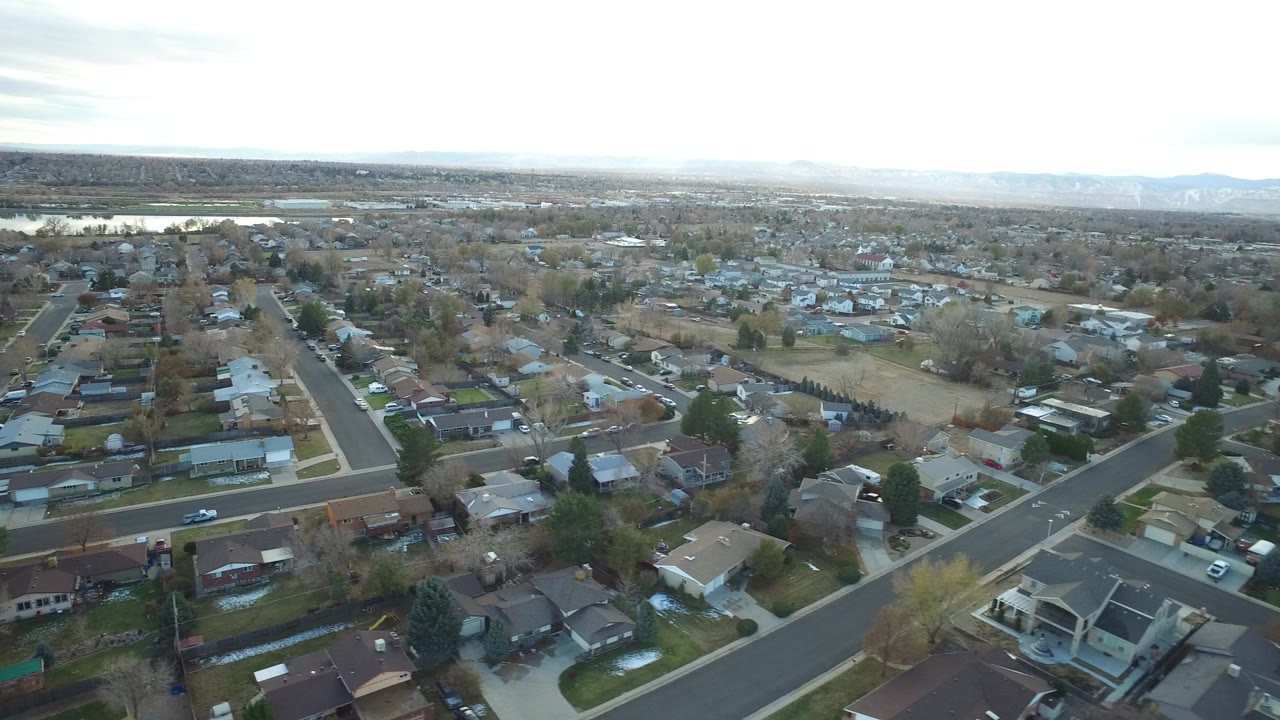 Wide aerial frame over Westminster, Colorado homes and street corners with pale sky, distant hills, and no visible people.
