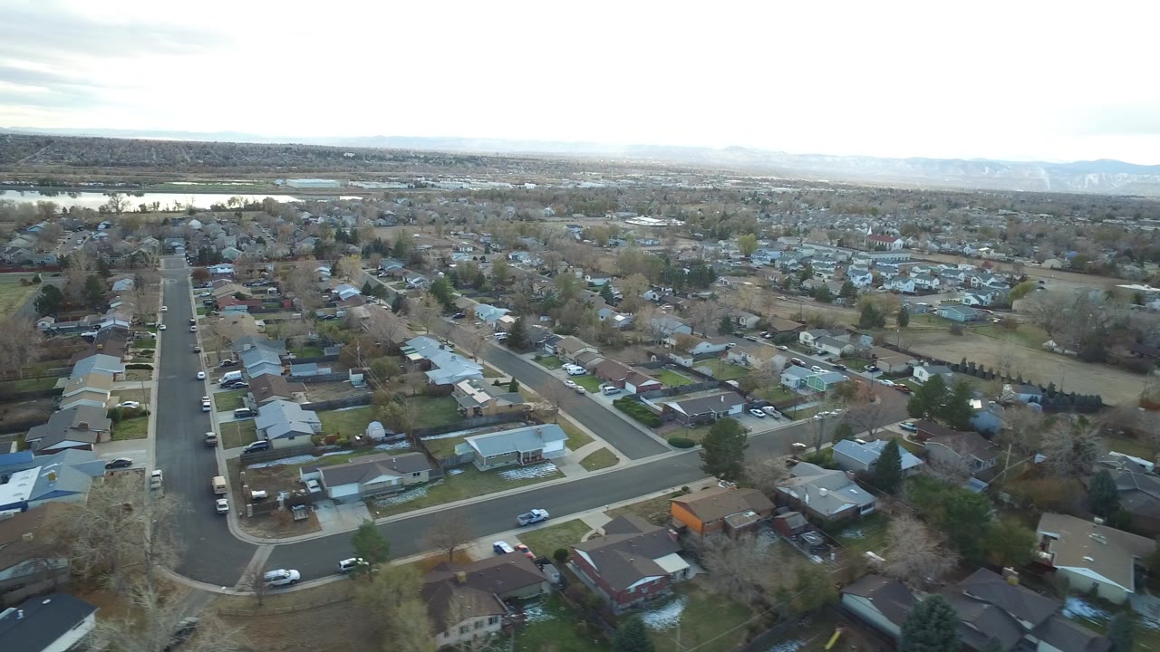 Aerial view across Westminster, Colorado residential blocks, with a straight street in the foreground, rows of houses, leafless trees, distant lake water, and no visible people.