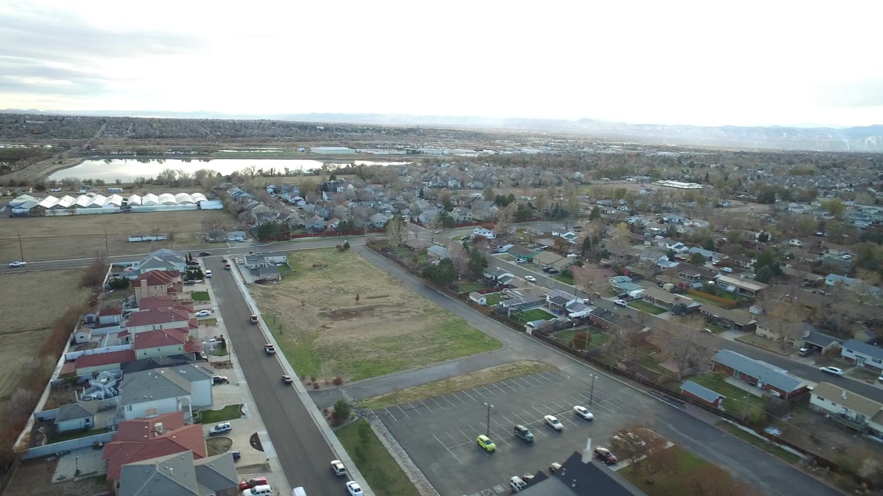 Aerial view over houses, streets, fields, and a distant water body.