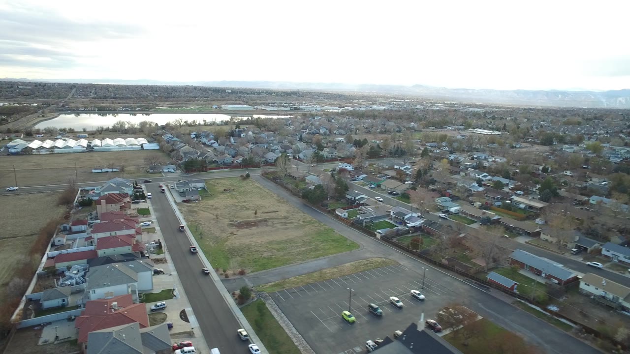 Westminster, Colorado aerial frame with red and gray rooftops in the foreground, an empty parking lot, neighborhood streets, fields, distant water, and no visible people.