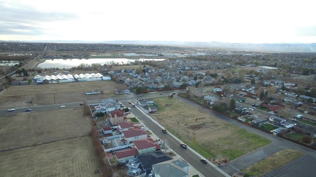 Aerial view of suburban homes, fields, roads, distant water, and cloudy sky.