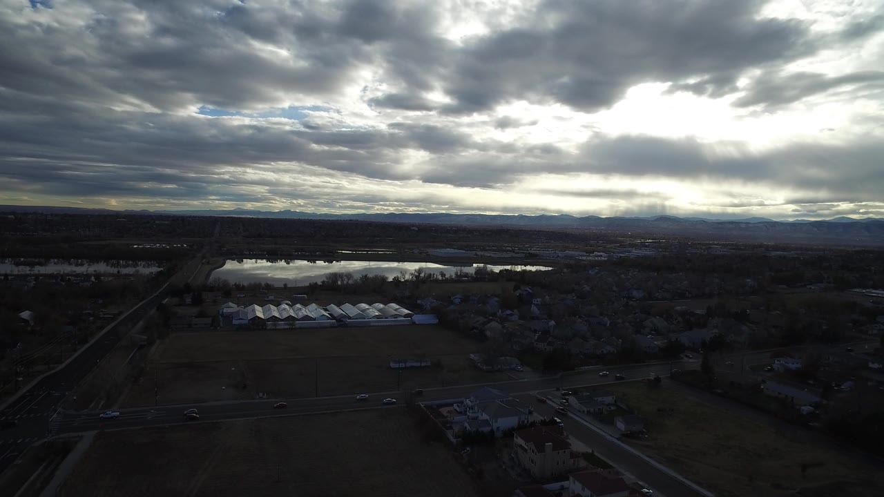 Aerial view over roads, fields, low buildings, water, and heavy clouds.