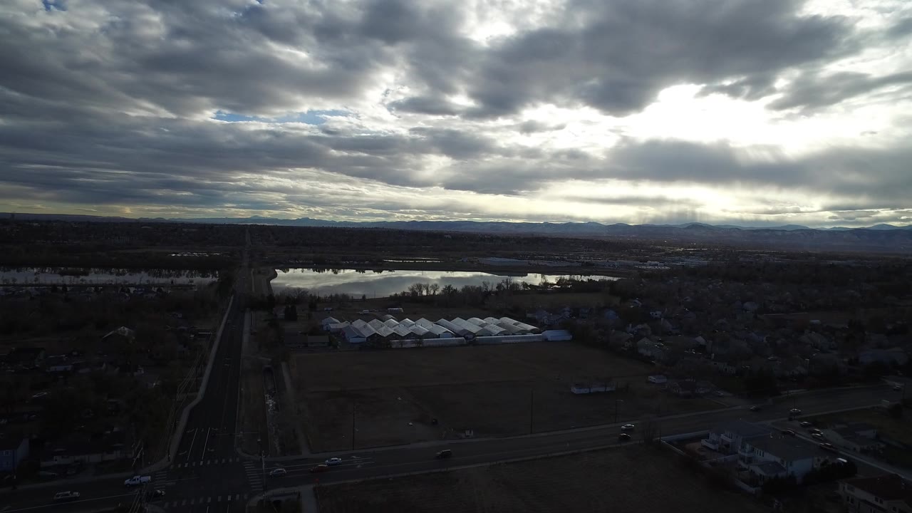 Opening aerial frame over Westminster, Colorado with dark fields, a road crossing the lower edge, reflective water beyond the fields, distant mountains, heavy clouds, and no visible people.