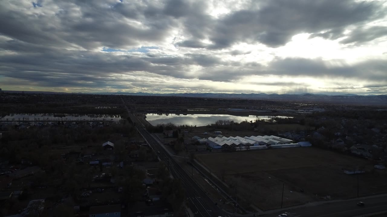 Second aerial frame over Westminster, Colorado showing the water catching pale sky light, dark tree cover, a straight road, fields, low structures, and no visible people.