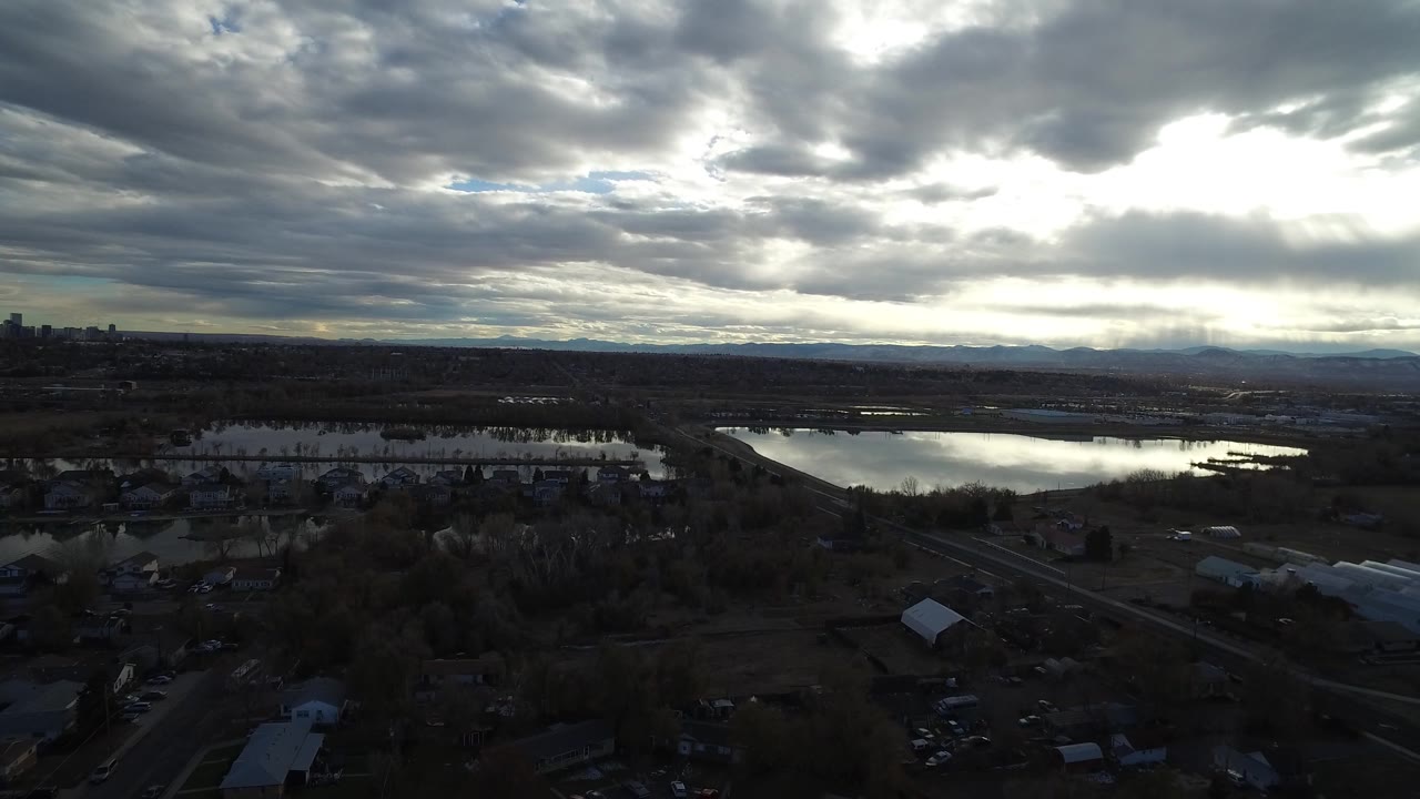 Aerial frame over Westminster, Colorado showing reflective wetland pools across the center, suburban structures below, distant low mountains, bright cloud breaks, and no visible people.