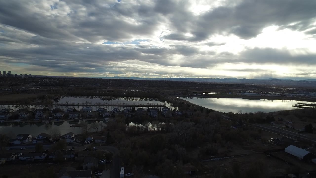 A slightly shifted wide frame shows reflective water cutting through Westminster marshland, with dark rooftops below and a distant skyline at the right, no visible people.