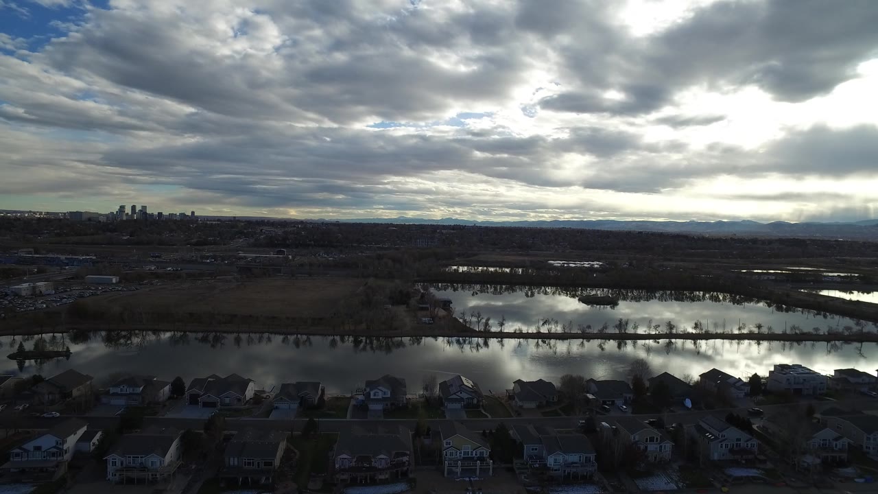Elevated Westminster, Colorado frame with dark residential rooftops in the foreground, a band of floodwater in the midground, bright cloud reflections, and no visible people.