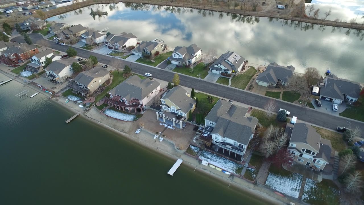 A slightly shifted aerial view in Westminster, Colorado shows shoreline houses, a paved road, small docks, reflective water, lawns, and no visible people.