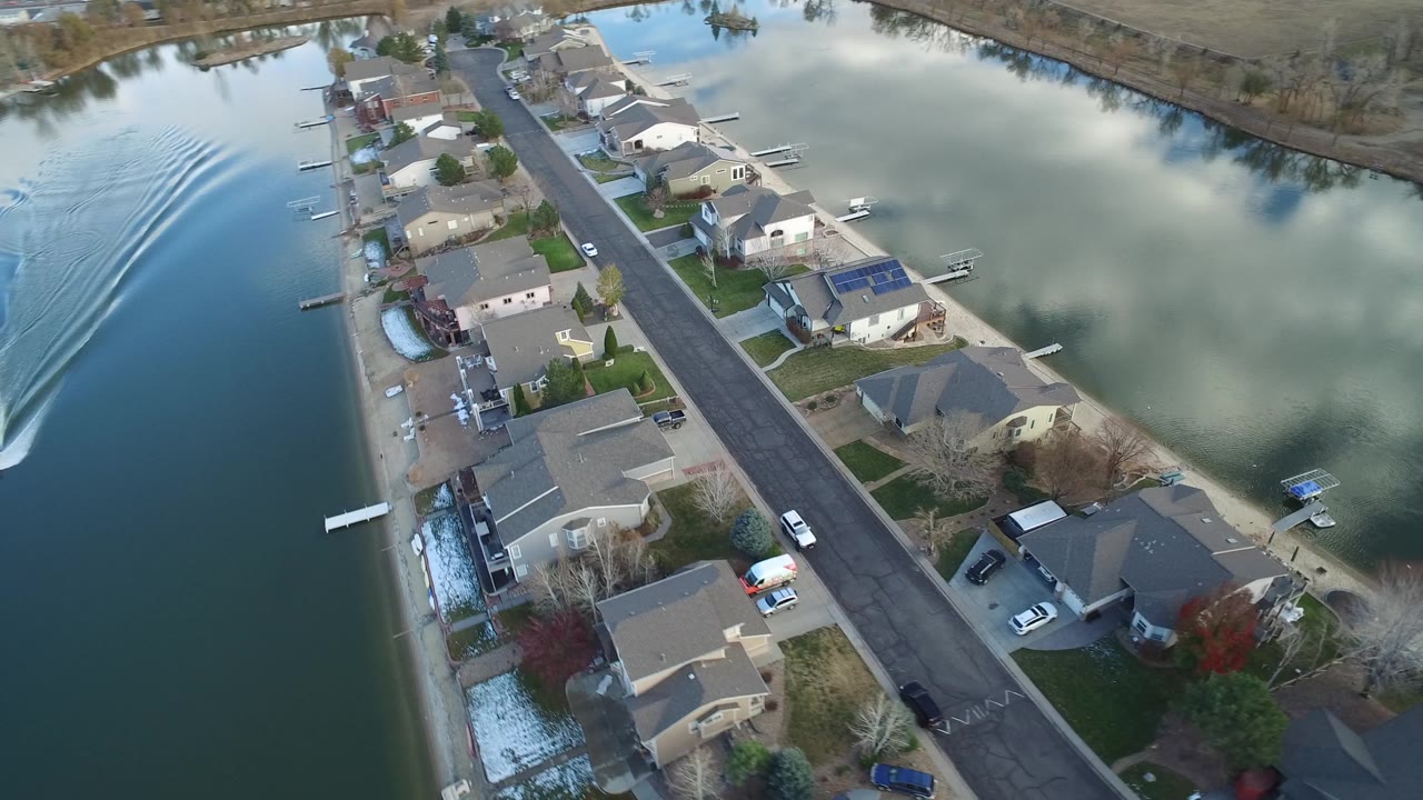 Top-down Westminster, Colorado neighborhood view with homes, docks, parked cars, reflected clouds, and a boat wake spreading wider across the left waterway with no visible people.