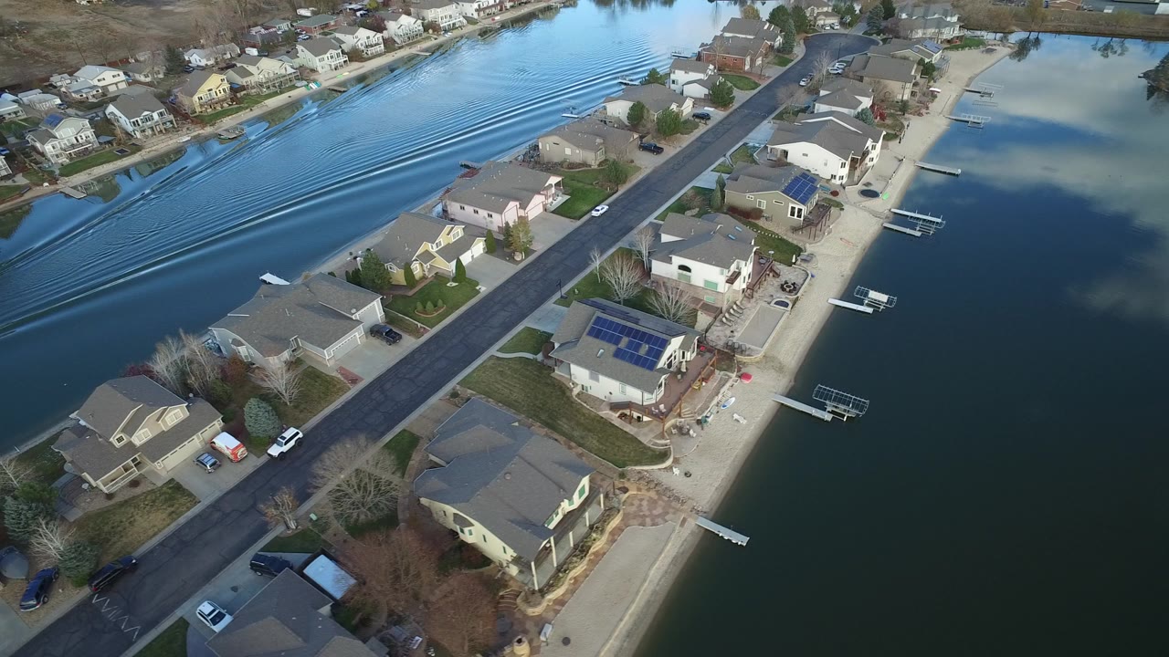 A high aerial frame over Westminster, Colorado shows waterfront houses, a street, private docks, and long wake lines crossing the channel, with tiny people visible near the homes.