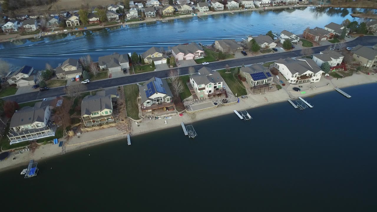 High oblique frame over Westminster, Colorado showing more of the dock line, sandy waterfront, large houses, residential street, and blue waterway beyond the homes.