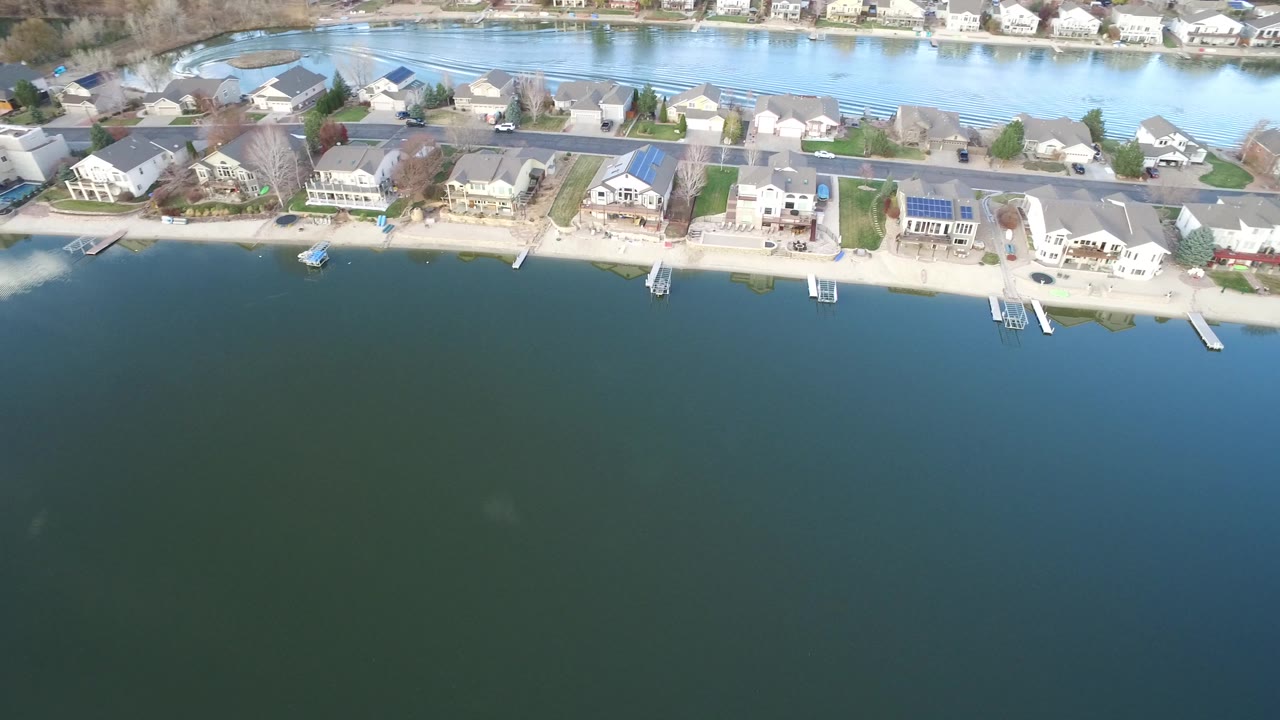 The aerial view continues over Westminster, Colorado with water across the lower frame, a pale bank, private homes, docks, and a road running behind the houses, with no visible people.