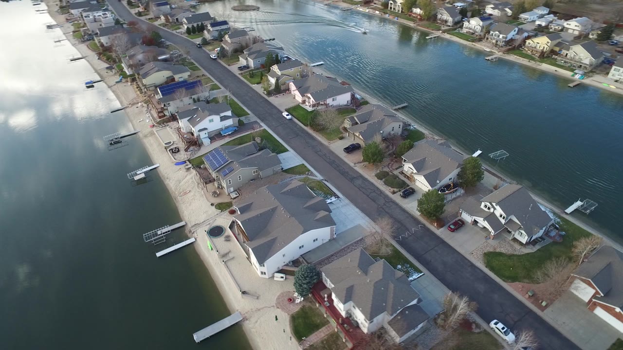 Aerial view in Westminster, Colorado showing private homes beside a narrow waterway, with docks, a road, driveways, and cloud reflections on the water.
