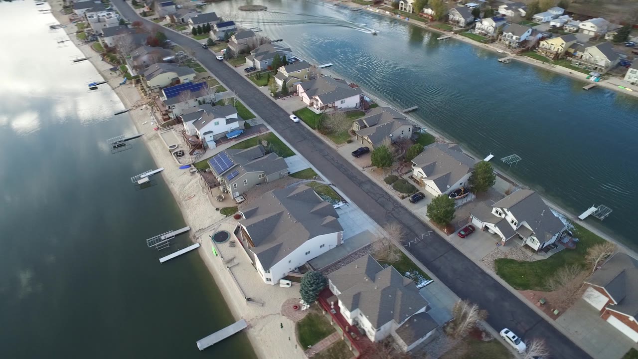Aerial frame in Westminster, Colorado with houses and parked vehicles along a straight residential street, docks on both banks, and bright cloud reflections across the water.