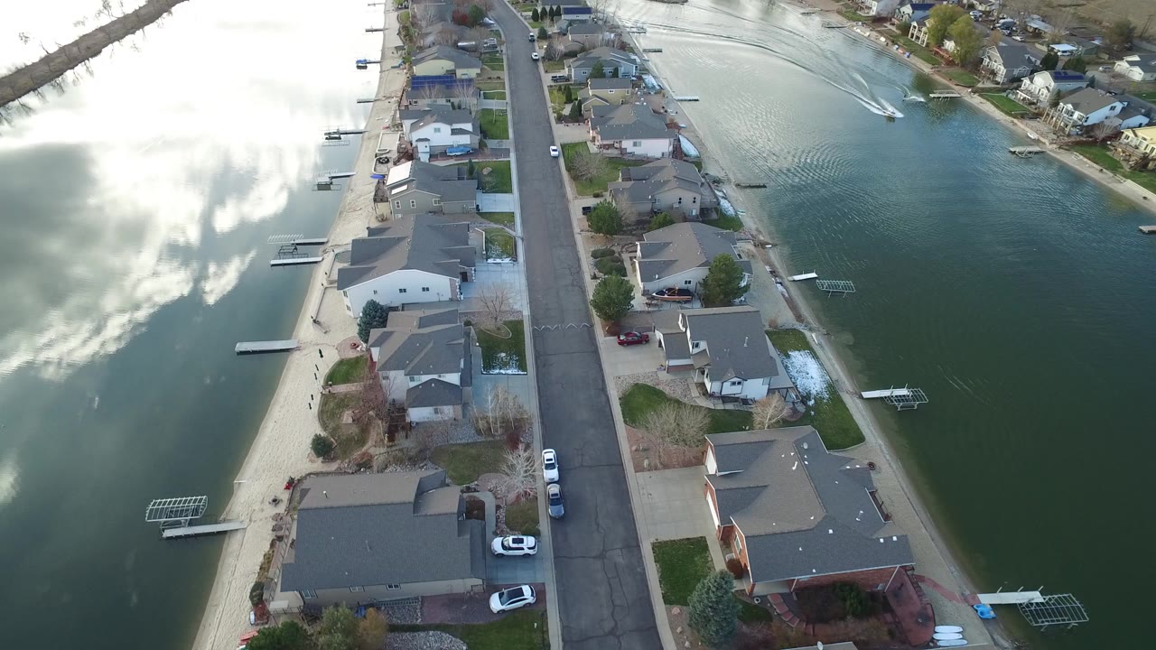 Aerial view over Westminster, Colorado homes beside a narrow lake, with docks, parked cars, cloud reflections on the water, and no visible people.
