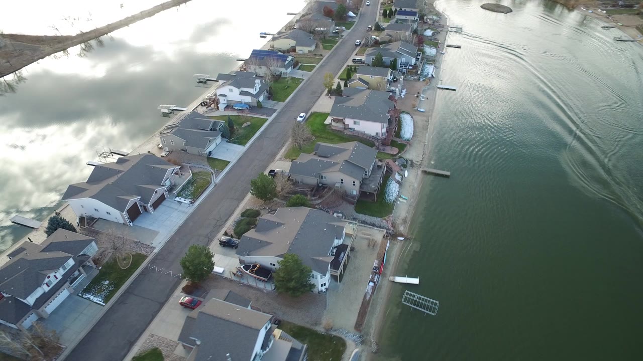 High-angle aerial view in Westminster, Colorado showing a row of private homes along a narrow road beside reflective green water, with docks and no visible people.