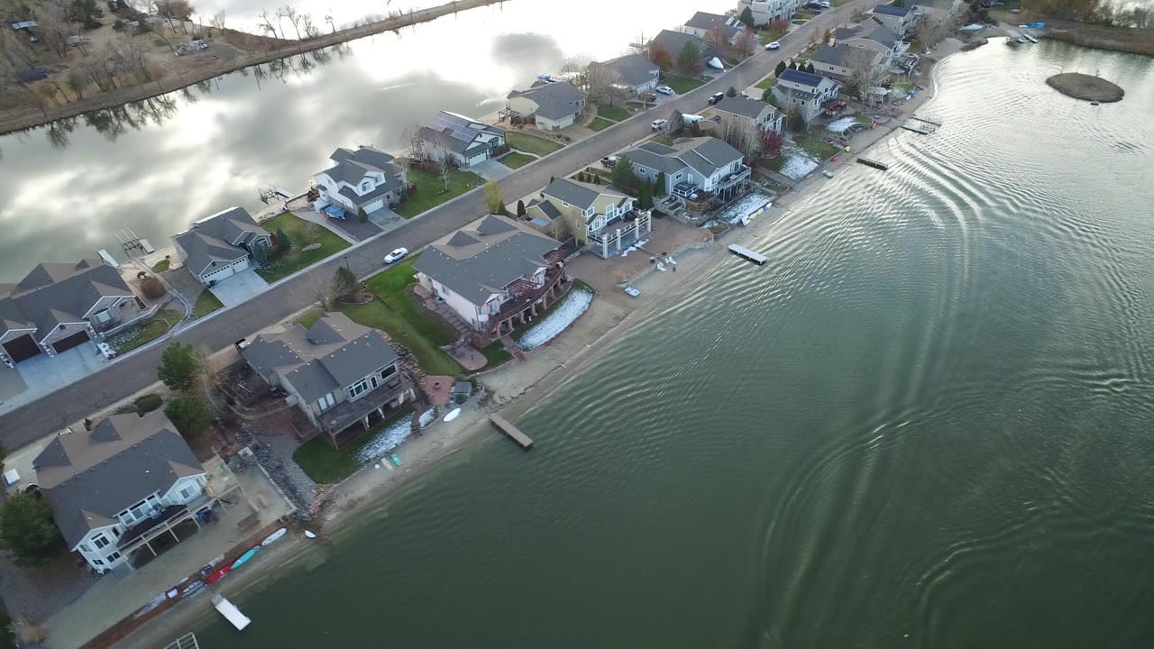 Aerial view in Westminster, Colorado with lakeside houses along the lower shore, a neighborhood road behind them, and curved wake lines spreading across green-gray water with no clearly visible people.