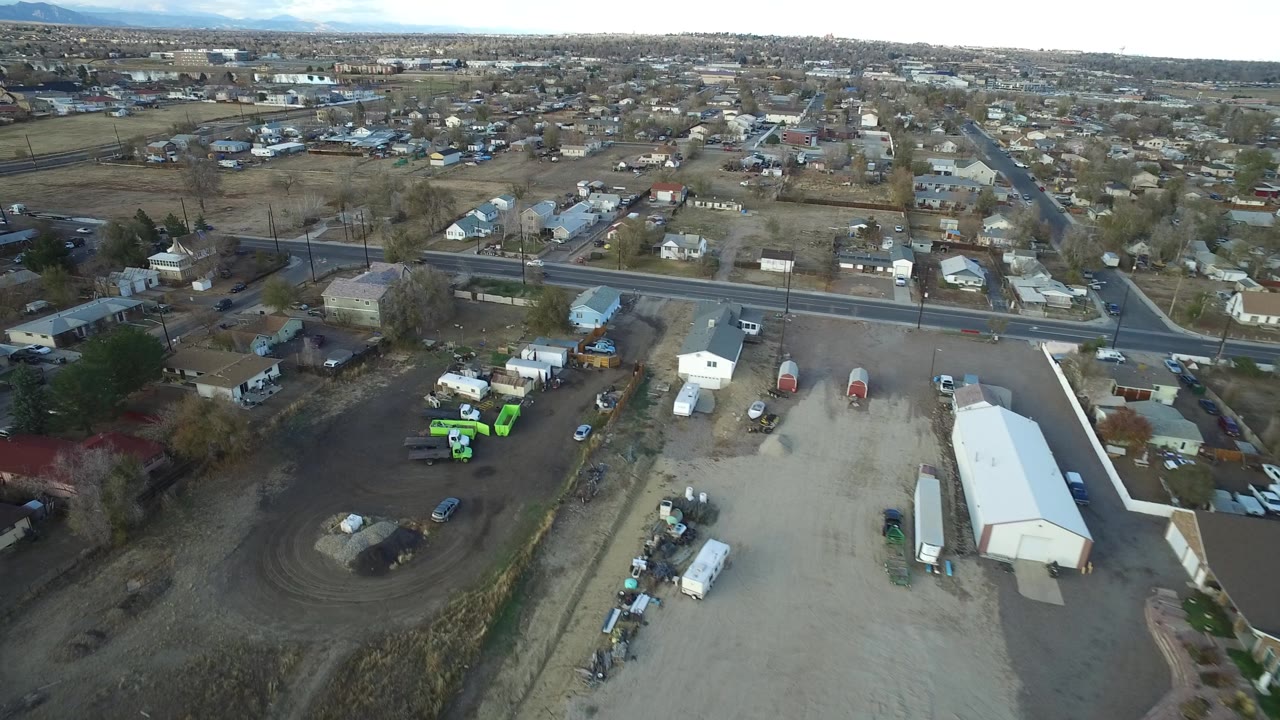 Aerial frame in Westminster, Colorado with a light-roofed warehouse in the foreground, a main road across the center, scattered houses beyond it, and no visible people.