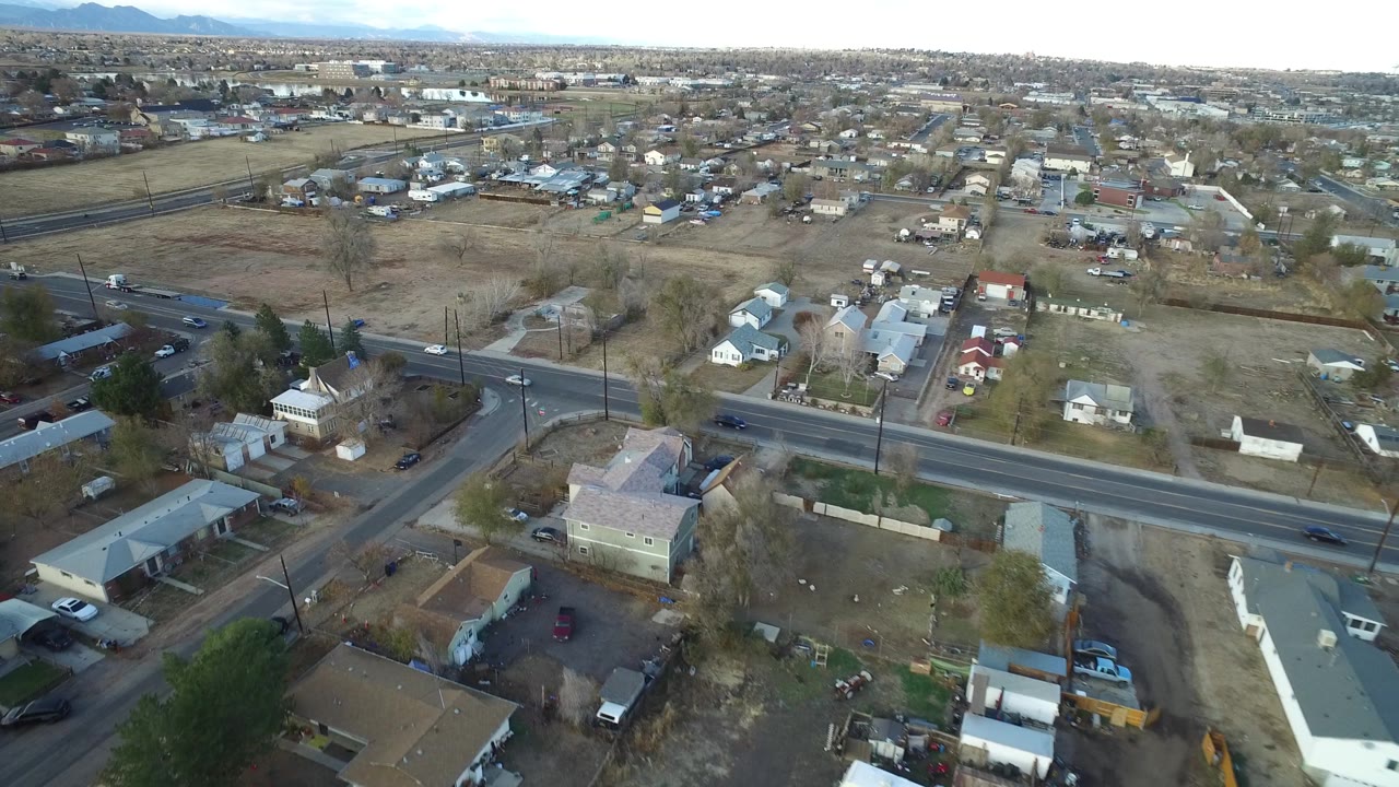 Drone view across Westminster, Colorado with a road crossing the foreground, low houses and yards below, open land in the midground, and no visible people.