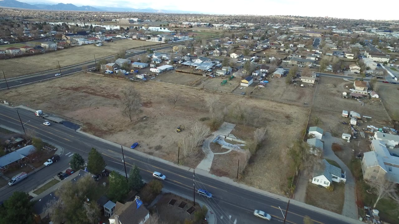 Drone-height view in Westminster, Colorado with a road crossing the foreground, pale open land, small houses and sheds in the midground, distant mountains, and no visible people.