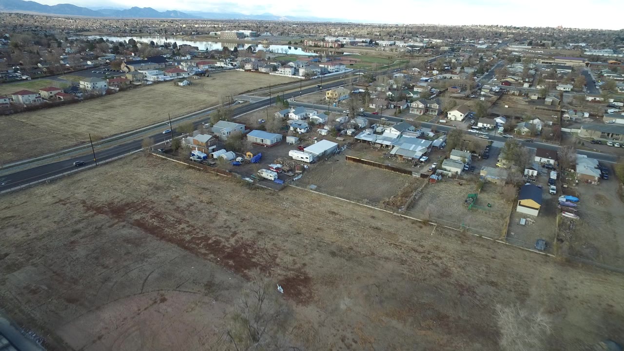 Aerial view over Westminster, Colorado with a dry open field in front, a road with cars, scattered houses, distant water, mountains on the horizon, and no visible people.
