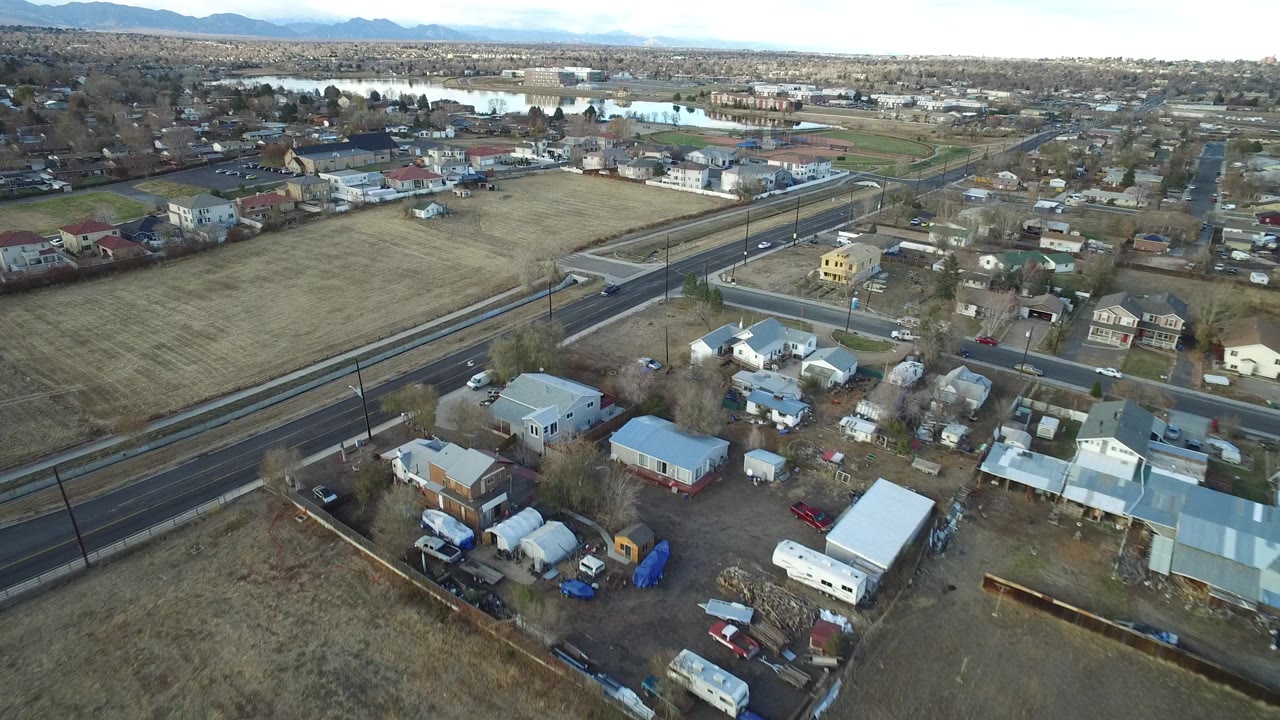 Aerial view in Westminster, Colorado looking across roadside homes, parked vehicles, a tan open lot, distant pond or lake, low mountains, and no visible people.