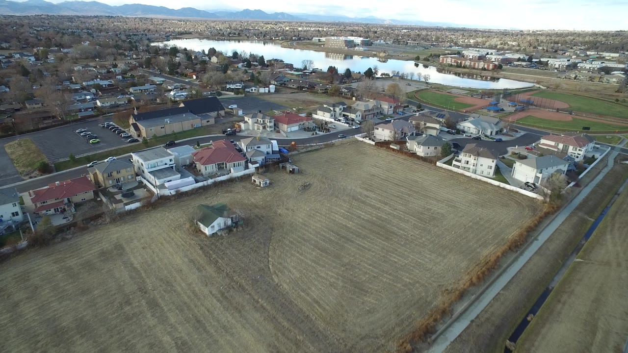 The next aerial frame in Westminster, Colorado centers homes and a church-like building near a large field, with the lake and mountain horizon behind them and no visible people.