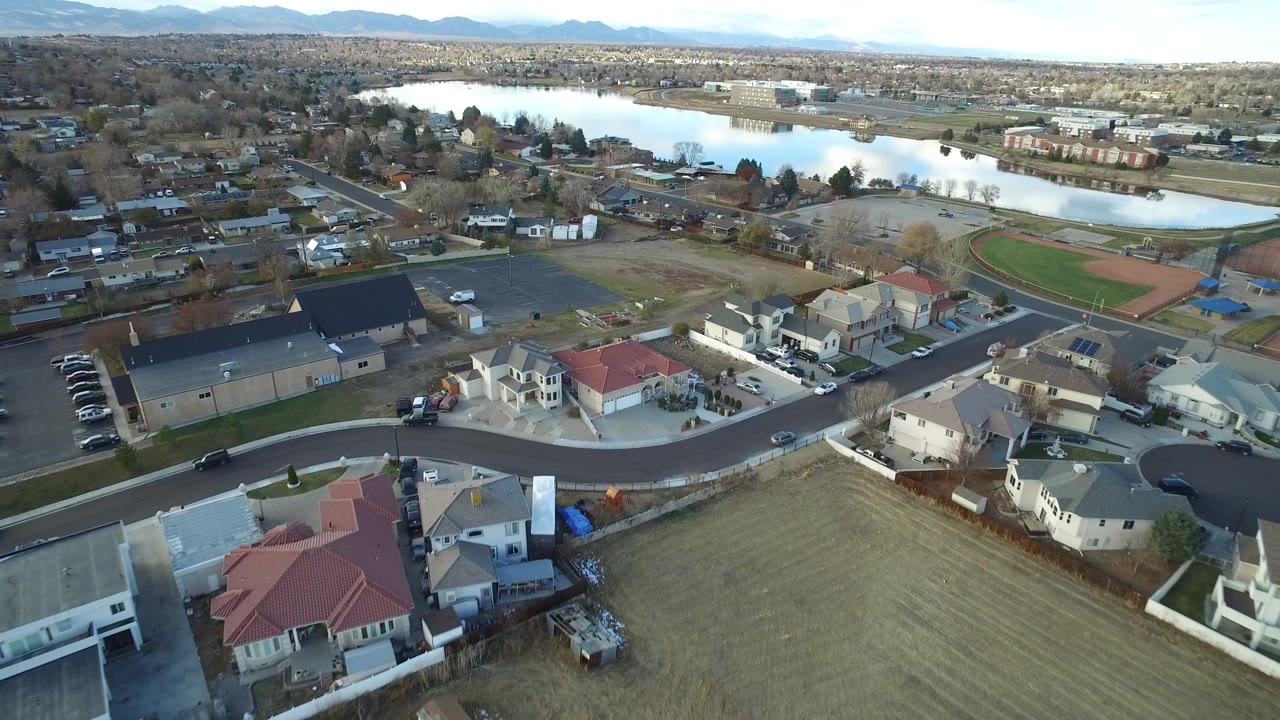 Aerial view in Westminster, Colorado with a large dark-roofed building and parking lot near the center, red-roofed homes below, a baseball field to the right, and no visible people.