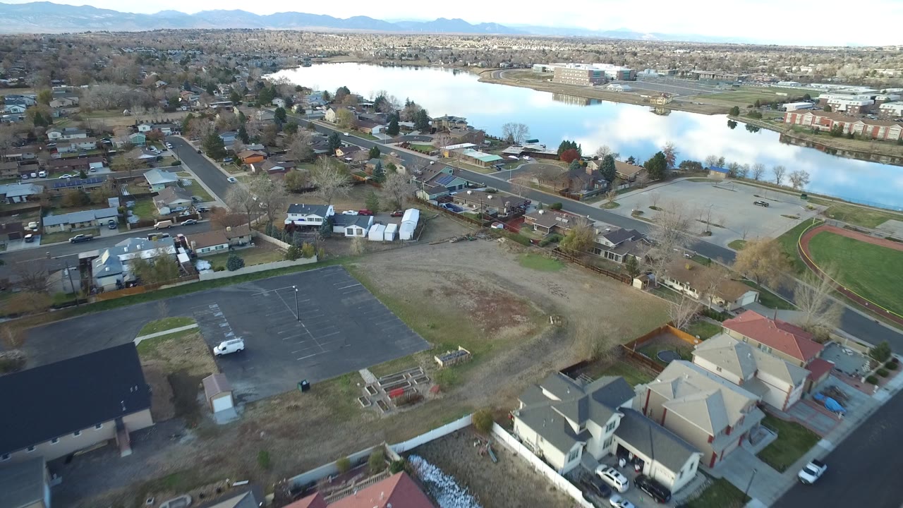 Aerial frame over Westminster, Colorado showing foreground rooftops, a mostly empty parking lot, neighborhood blocks, and a broad lake reflecting the pale sky.