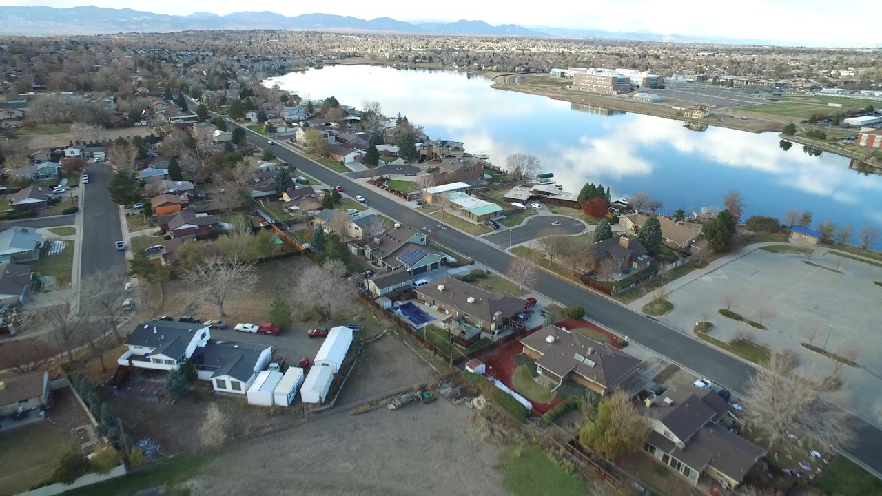 Aerial view over Westminster, Colorado with residential streets and houses in the foreground, a reflective lake beyond them, distant mountains, and no visible people.