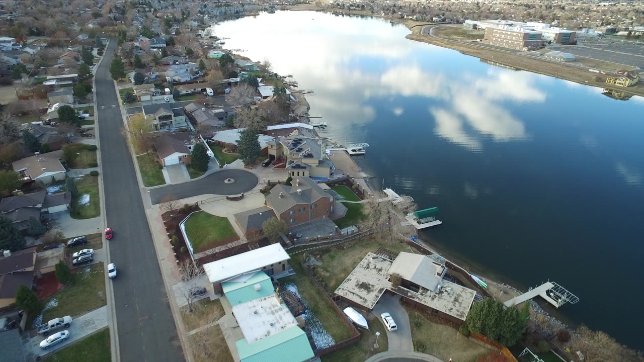 Aerial view over Westminster, Colorado lakefront homes, a straight residential road, docks, cloud reflections on deep blue water, and no visible people.