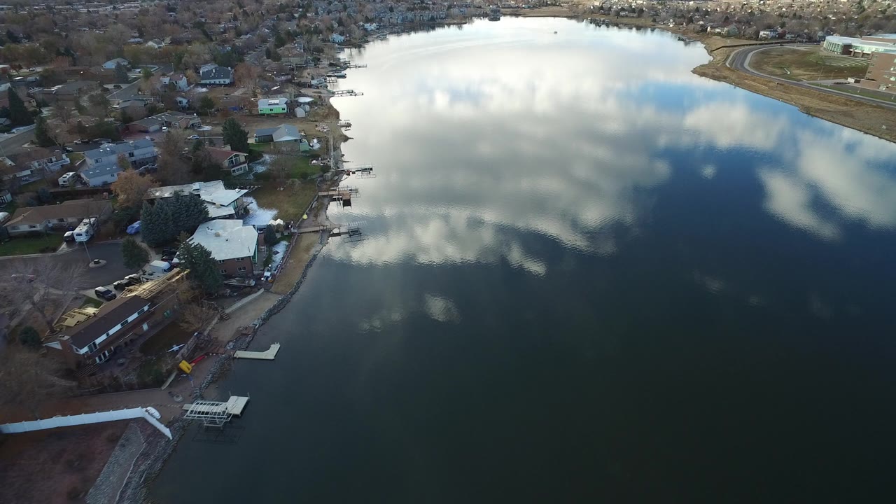 Aerial view in Westminster, Colorado showing a lake with cloud reflections, shoreline homes, small docks, bare trees, and no visible people.