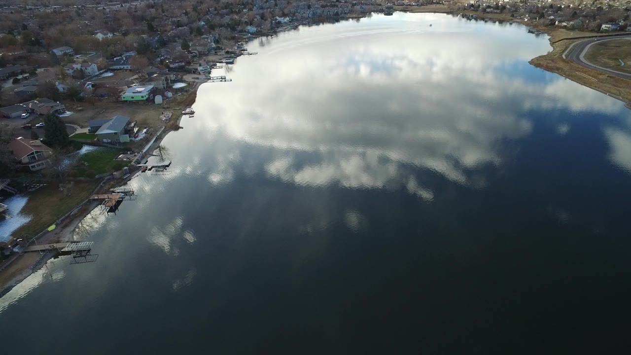 A high aerial frame in Westminster, Colorado shows bright clouds mirrored on the lake, with houses, yards, and a dock tracing the left shoreline, no visible people.