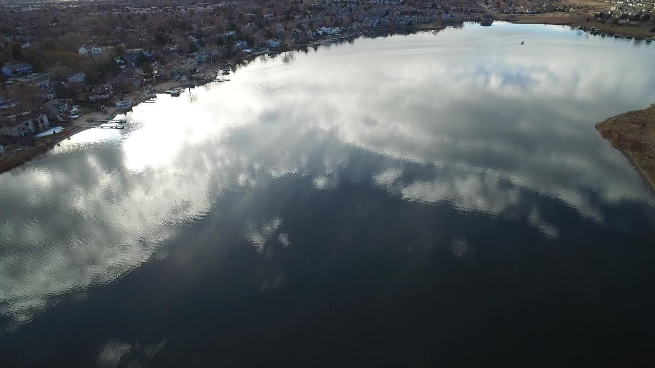 Aerial view over a dark reflective pond in Westminster, Colorado, with bright cloud reflections, a curved shoreline, waterside structures, and no visible people.
