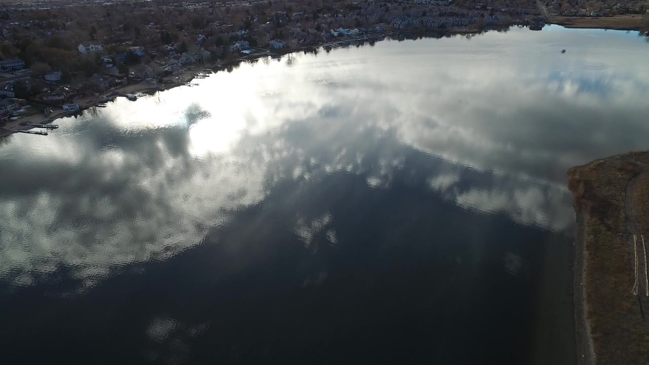 Aerial view of a lake reflecting bright clouds with shoreline homes at the top.