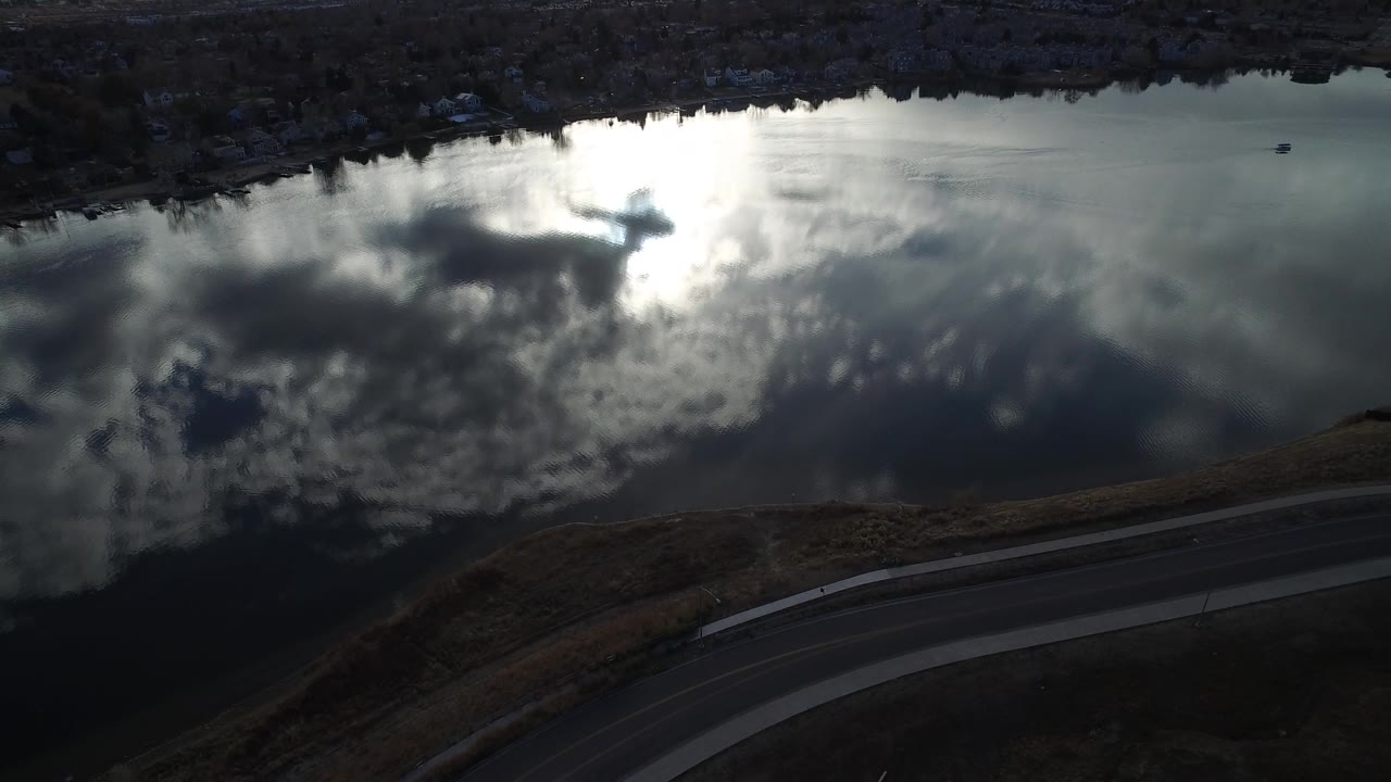 Aerial view of dark water reflecting bright clouds beside a curved road.