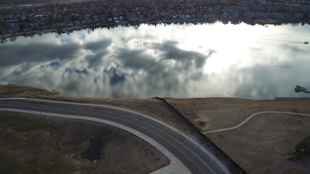 Opening aerial frame in Westminster, Colorado, showing bright cloud reflections on the water above a curved road, dry fields, small ponds, and no visible people.