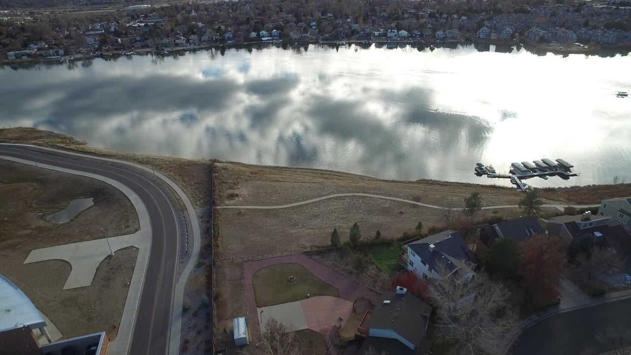 Aerial frame over Westminster, Colorado showing a reflective lake, shoreline docks, a curving road, private homes, winter lawns, and no visible people.