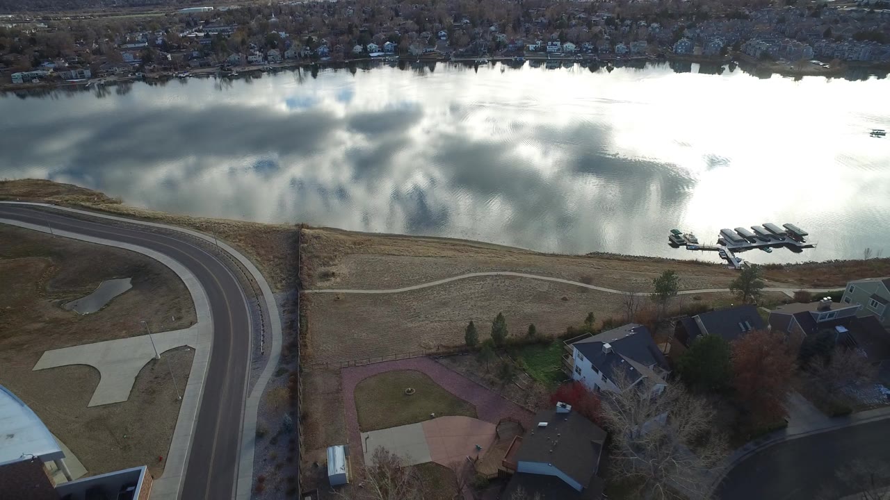 Aerial frame in Westminster, Colorado with bright cloud reflections on the lake, docks along the near shore, curved residential pavement, houses, and no visible people.