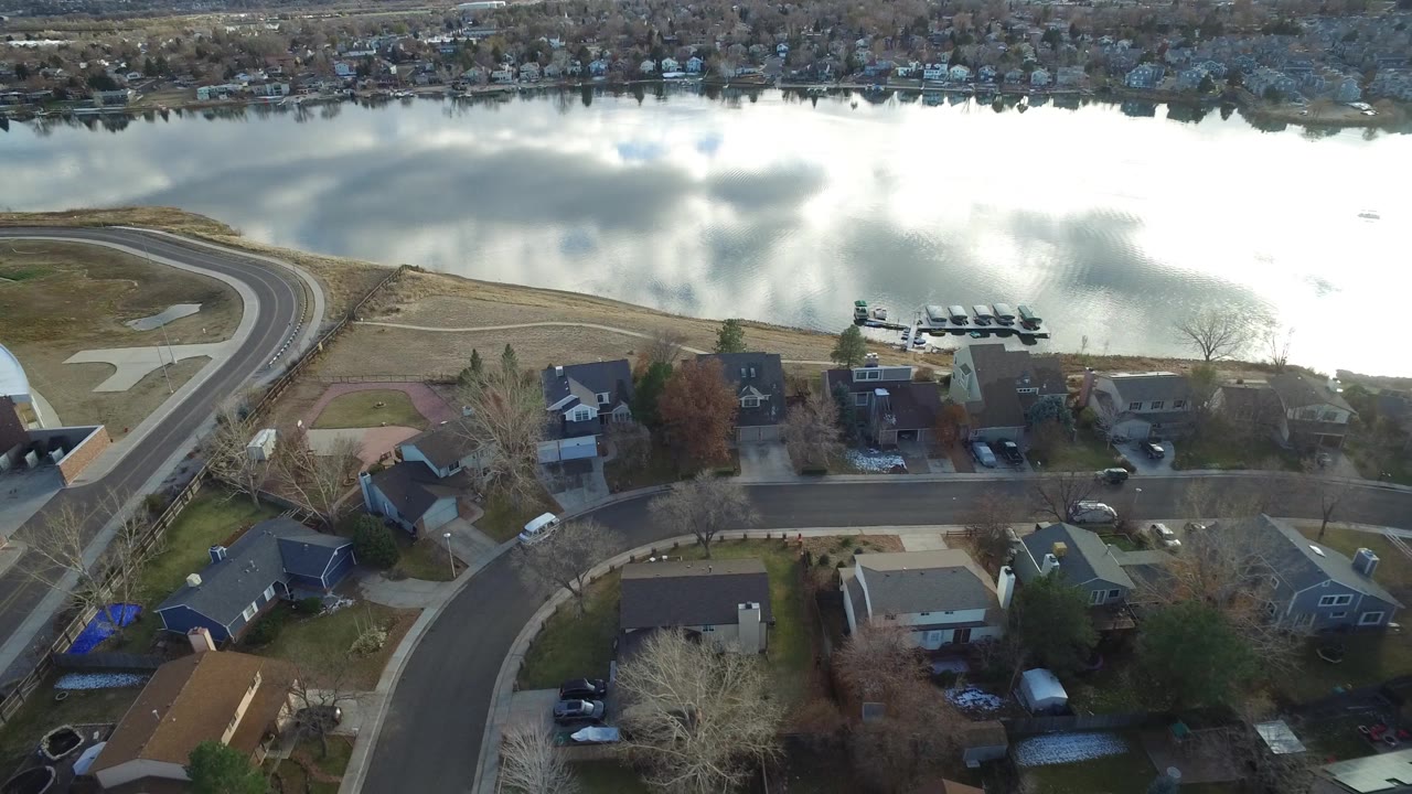 Aerial frame over a Westminster, Colorado residential block, with a curved street in the foreground, houses below, lake water behind, and no visible people.