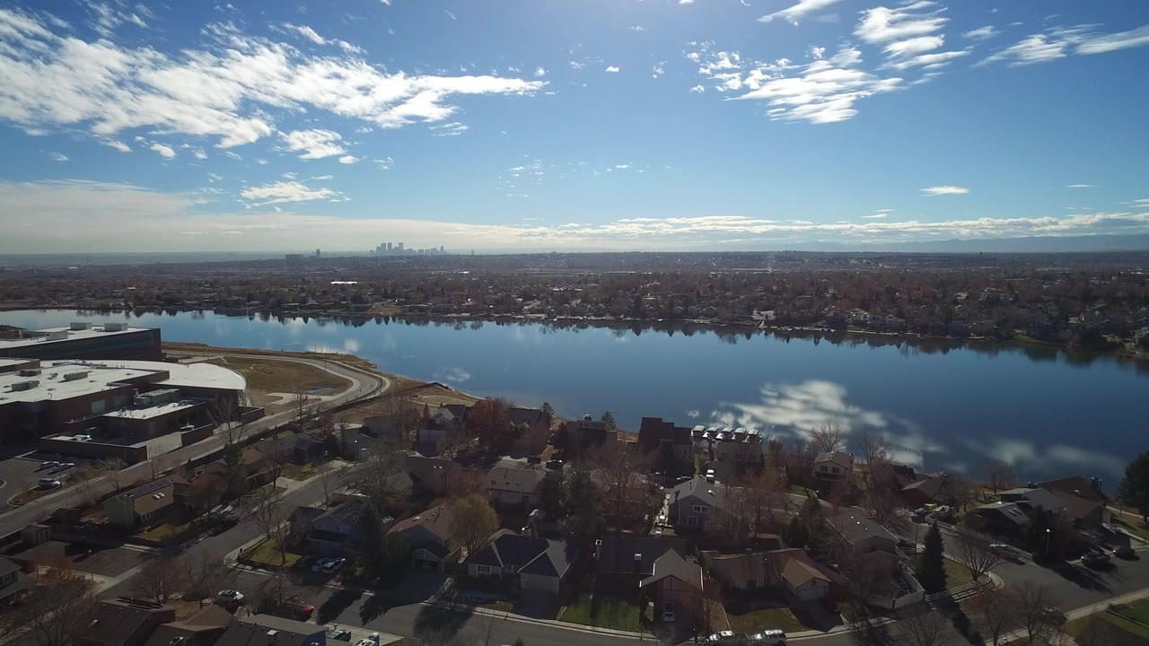 Wide drone view of a lake beside suburban streets and rooftops under a bright sky.