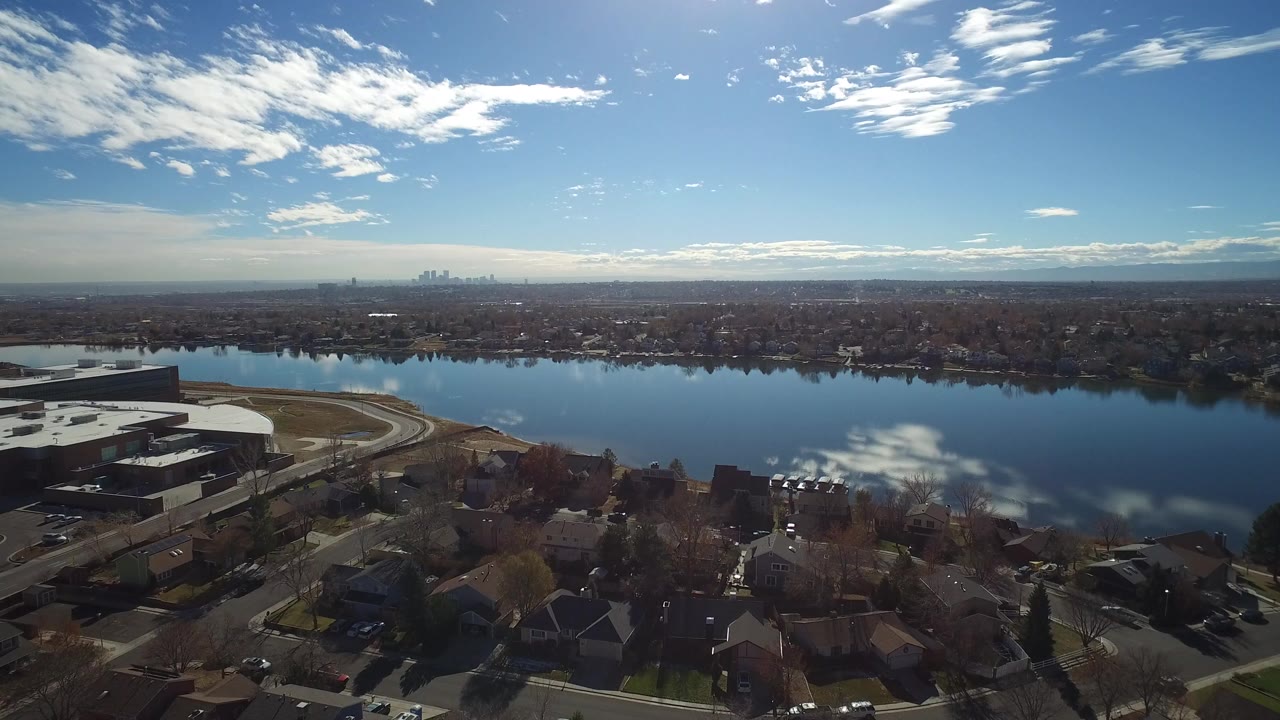 At 0:01, the Westminster aerial holds on the lake with residential blocks in the foreground, a reflective water surface, a far skyline, mountains, bright sky, and no visible people.